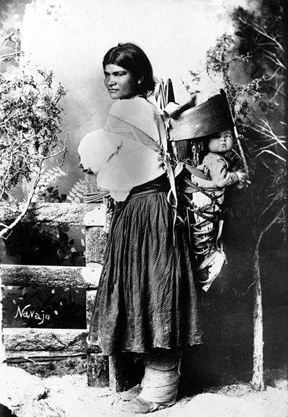 Retrato del estudio de una mujer nativa americana (Navajo) con un niño en una cuna portátil en su espalda. Ella lleva mocasines de arranque.